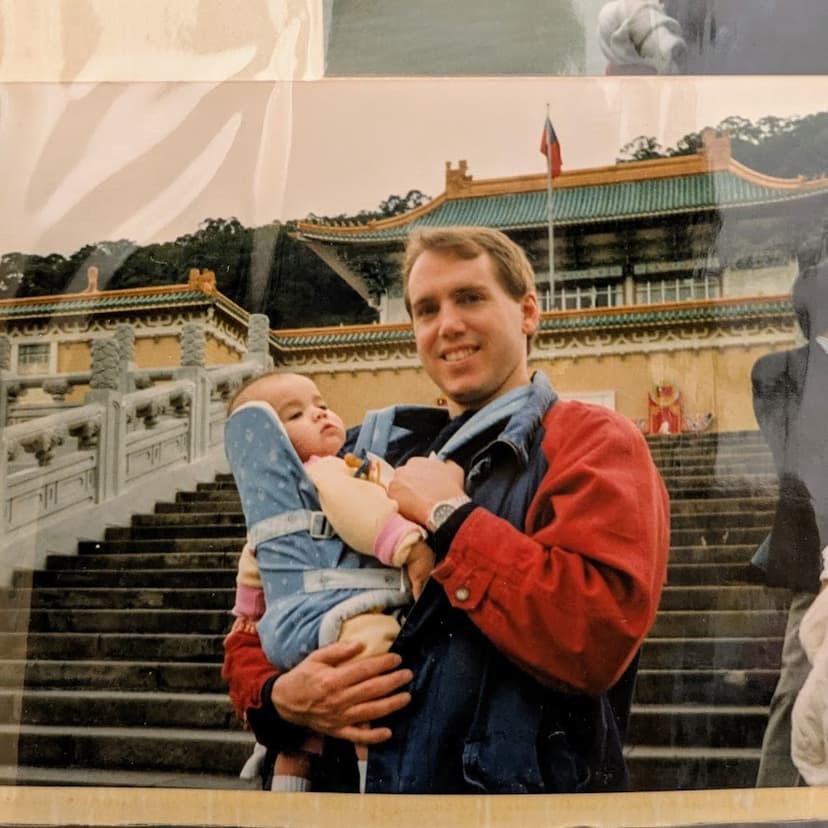 An old photograph of a man smiling and wearing a blue baby carrier with an infant on his front.