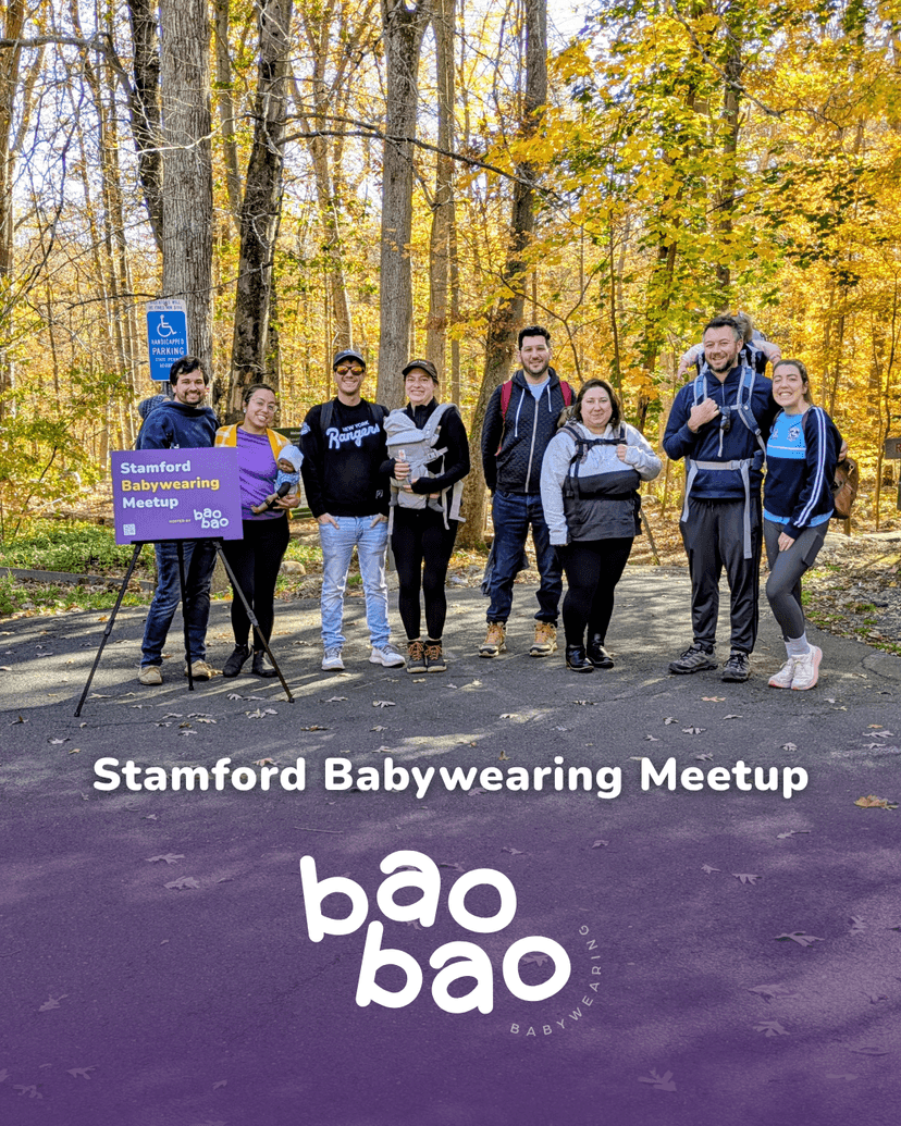 A group of four babywearing families outdoors smiling in front of brilliant fall foliage.