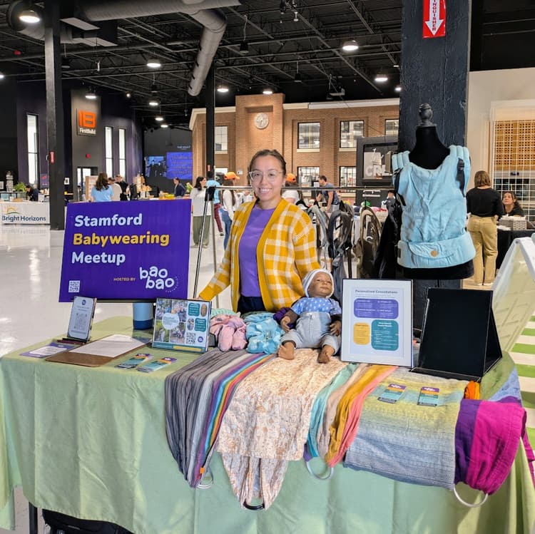 Waverly in an expo hall, at a table draped with carriers, dolls, and signs including Stamford Babywearing Meetup, surrounded by a rack of carriers and a mannequin wearing a carrier.