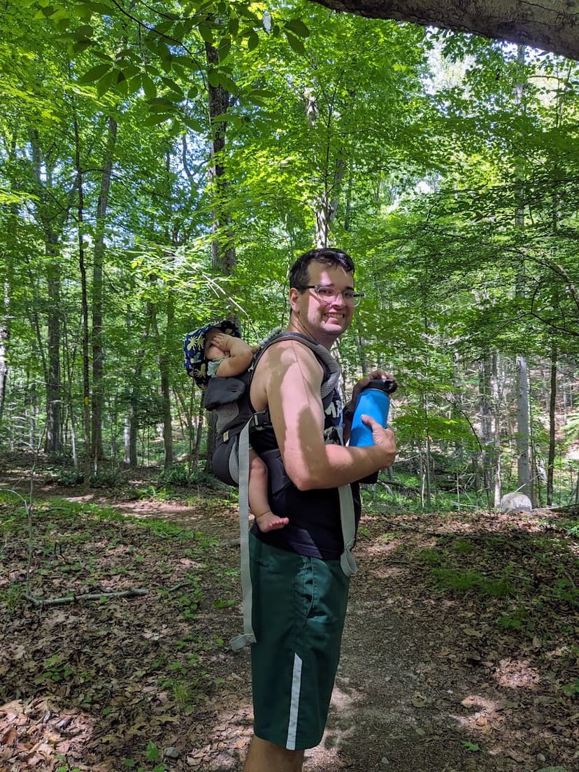 A man smiling on a forest path, wearing a soft-structured carrier with a child on his back.