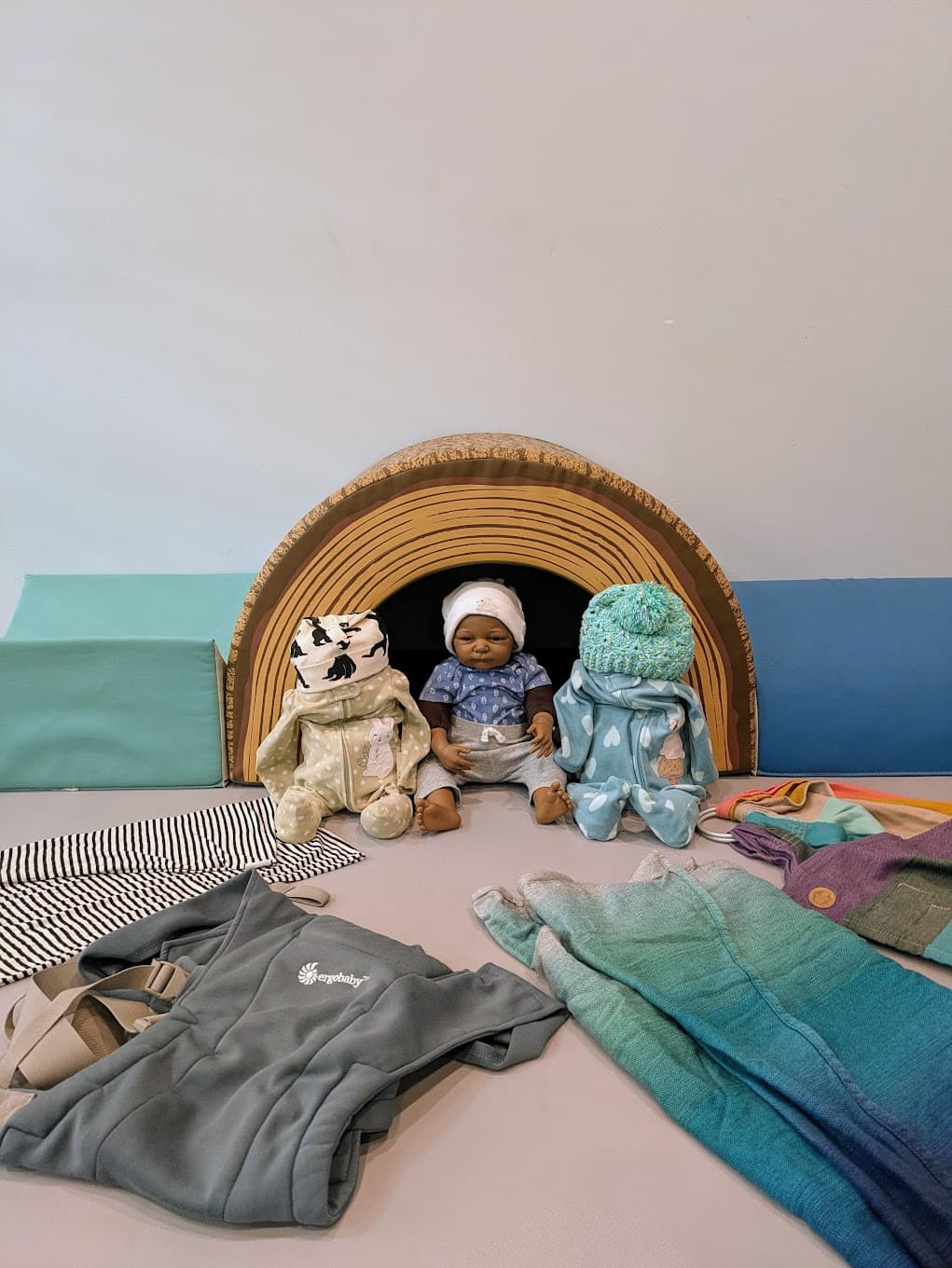 Three dolls sitting in front of soft play mats and surrounded by wraps and carriers.