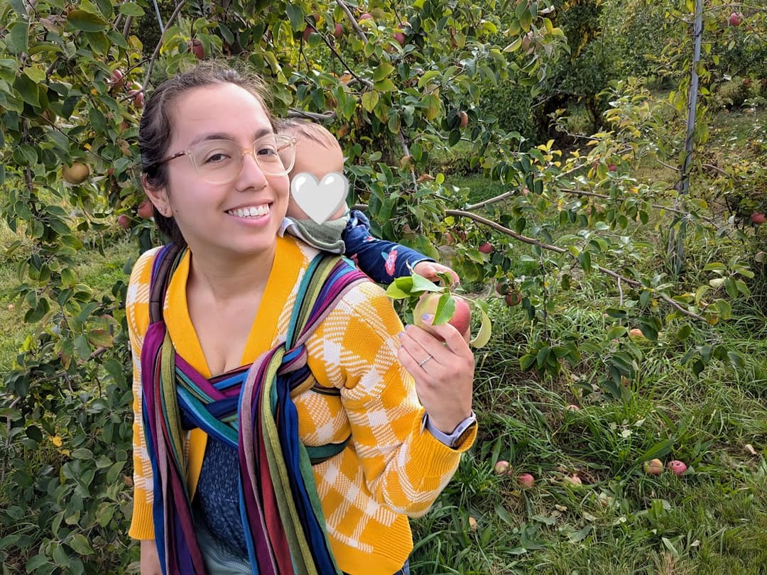 Waverly smiling, wearing a striped meh dai, handing an apple to a child on her back.