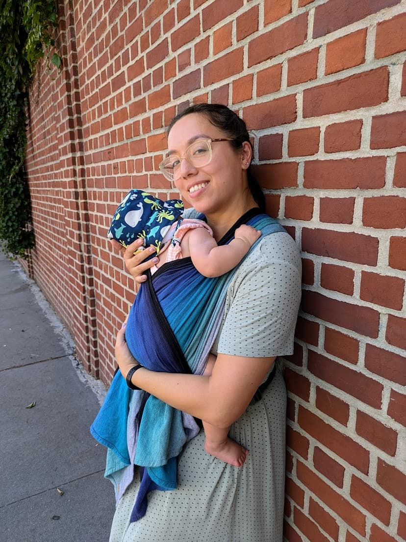 Waverly smiling and leaning againt a brick wall, wearing a blue woven wrap, snuggling a child on her front.